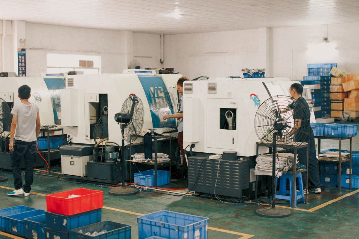 Workers operating CNC lathes on the floor of a small Chinese machine shop, with stacked plastic component crates and floor fans